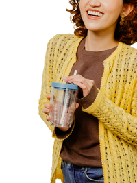A woman wearing a yellow open-front knit cardigan over a brown top, holding a clear insulated tumbler with a blue lid and straw against a white background.