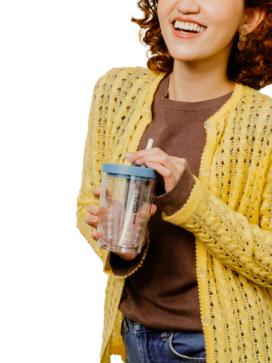 A woman wearing a yellow open-front knit cardigan over a brown top, holding a clear insulated tumbler with a blue lid and straw against a white background.