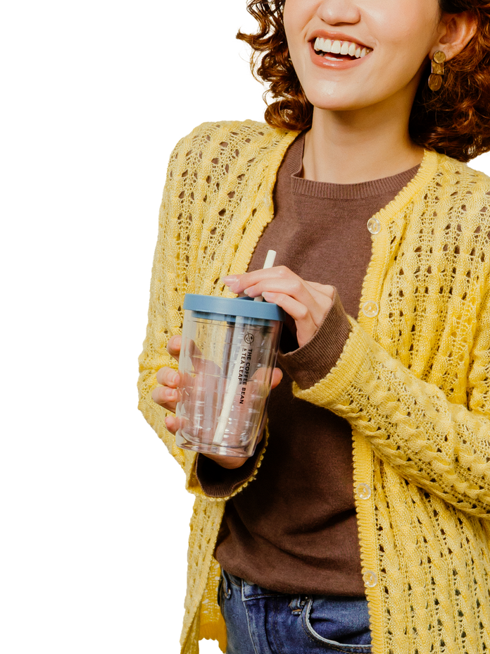 A woman wearing a yellow open-front knit cardigan over a brown top, holding a clear insulated tumbler with a blue lid and straw against a white background.