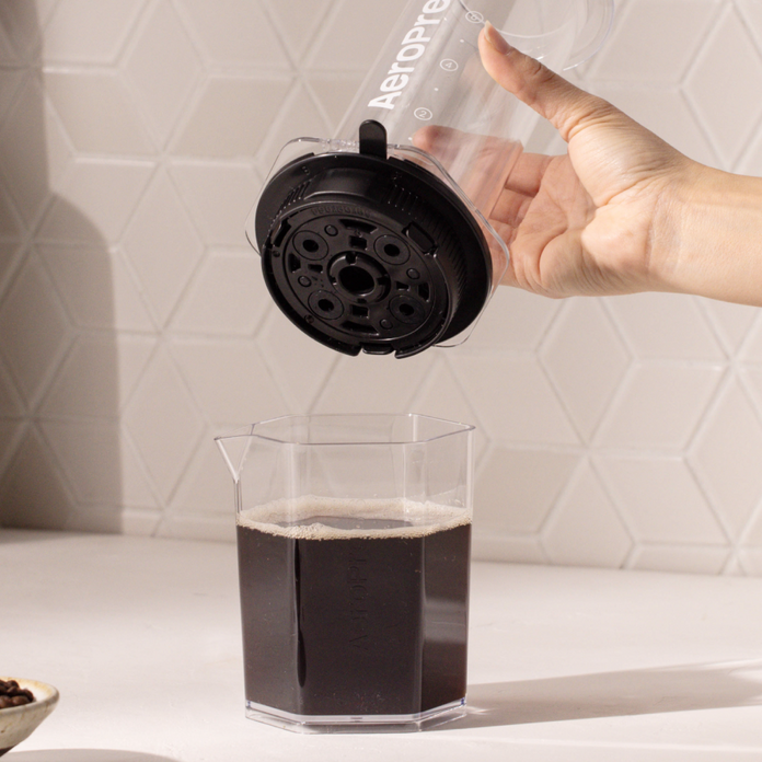 Hand pouring coffee from an AeroPress XL coffee press into a glass container on a white surface with a geometric tile pattern.