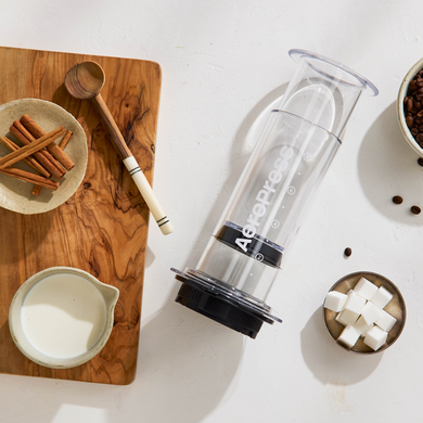 AeroPress XL coffee press on a white surface with wooden cutting board and ingredients.