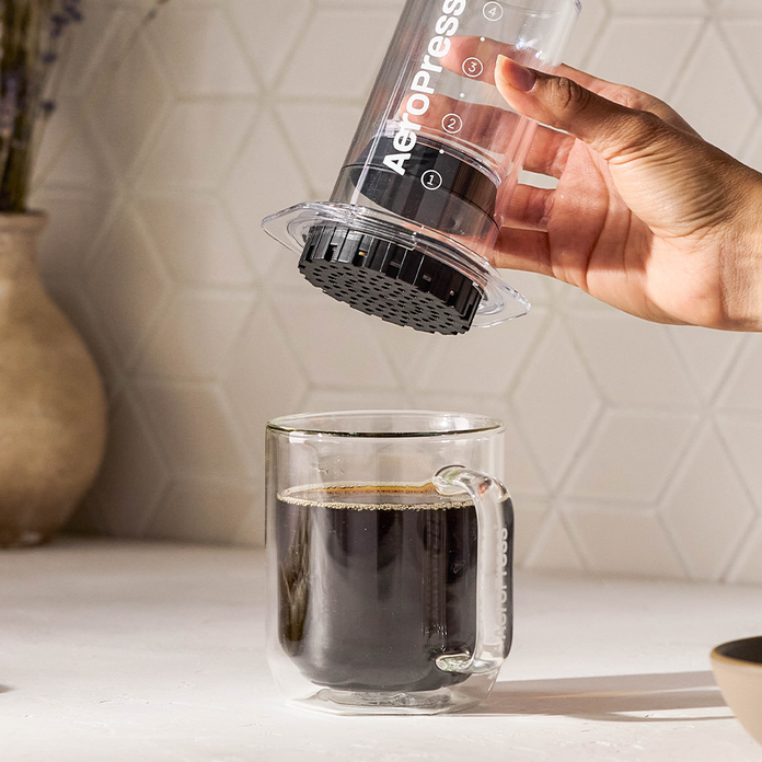 Hand using an Aeropress to pour coffee into a glass mug on a kitchen counter.