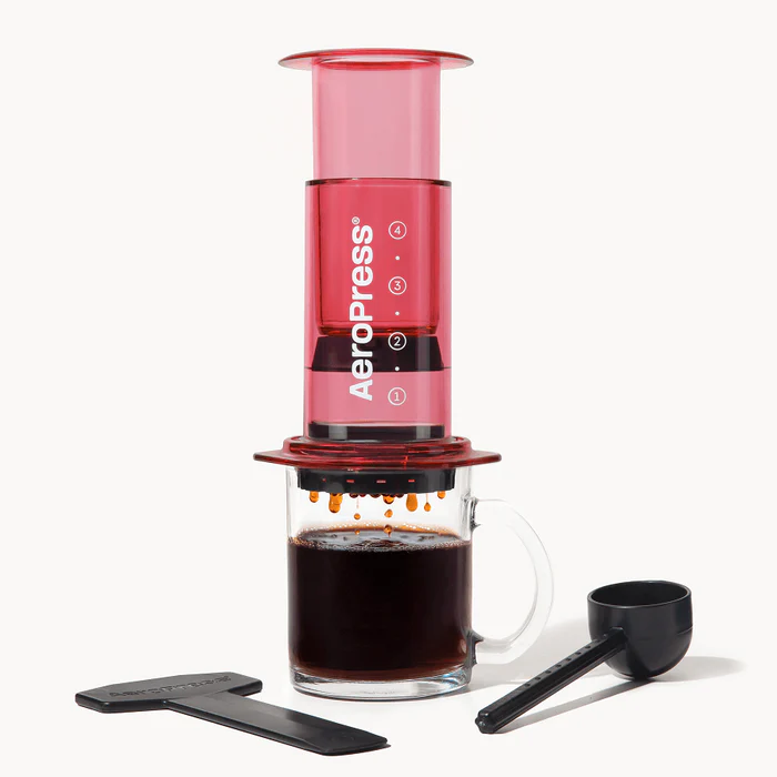 A coffee press from AeroPress in a translucent red, seeping coffee into through a filter into a clear coffee cup with accessories on each side.
