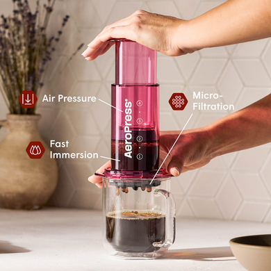 Person using a red Aeropress coffee maker with labeled features on a kitchen counter.