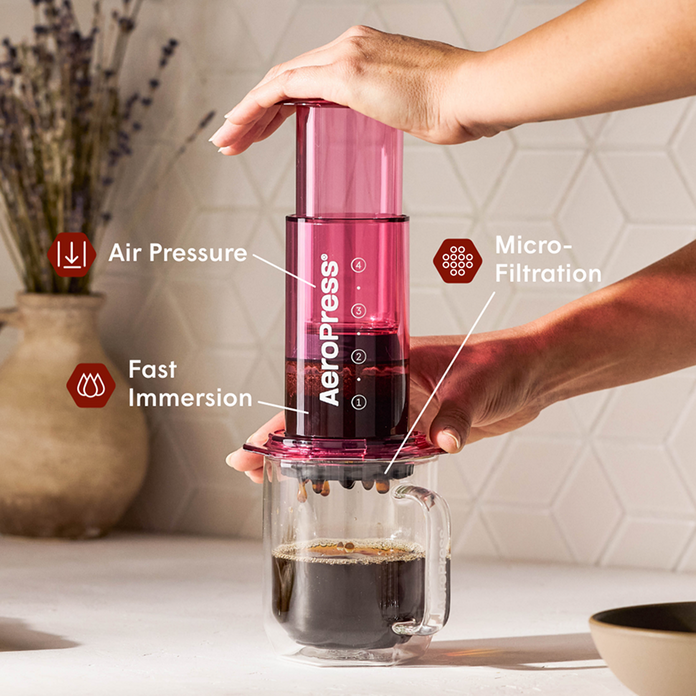Person using a red Aeropress coffee maker with labeled features on a kitchen counter.