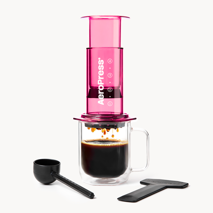 Pink AeroPress coffee maker with a glass mug and black scoop on a white background