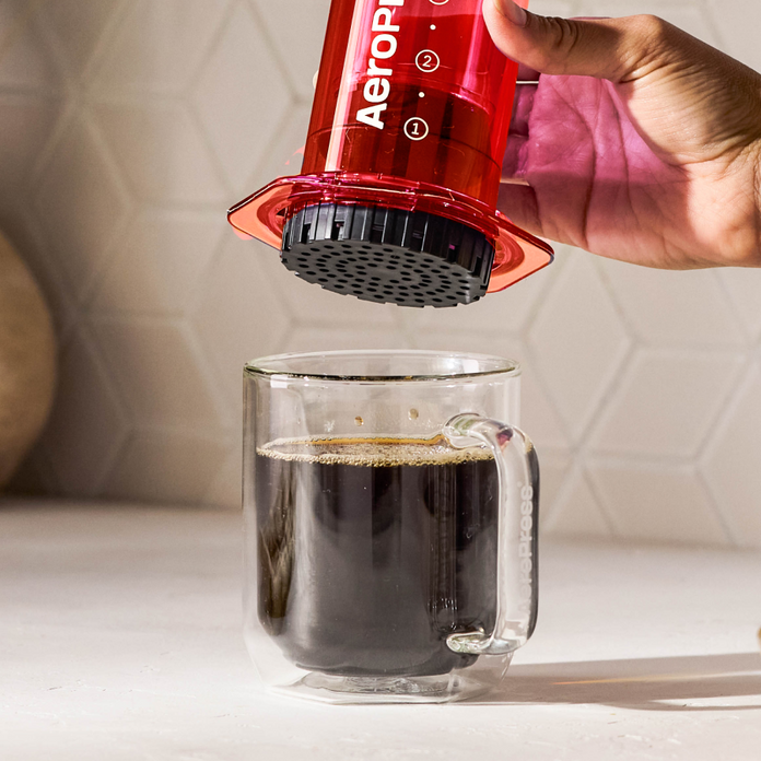 Red AeroPress device being used to make coffee in a clear glass mug.