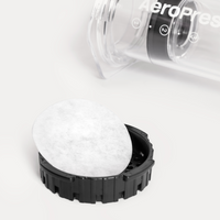 Close-up of a white paper filter in the lid of a coffee brewing device with a clear container and black base on a white background.