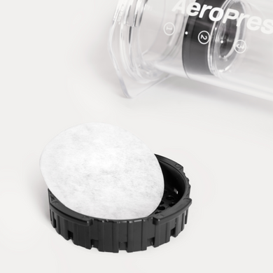 Close-up of a white paper filter in the lid of a coffee brewing device with a clear container and black base on a white background.