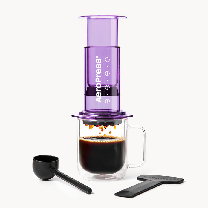 Purple AeroPress coffee maker with a glass mug of coffee and scoop on a white background