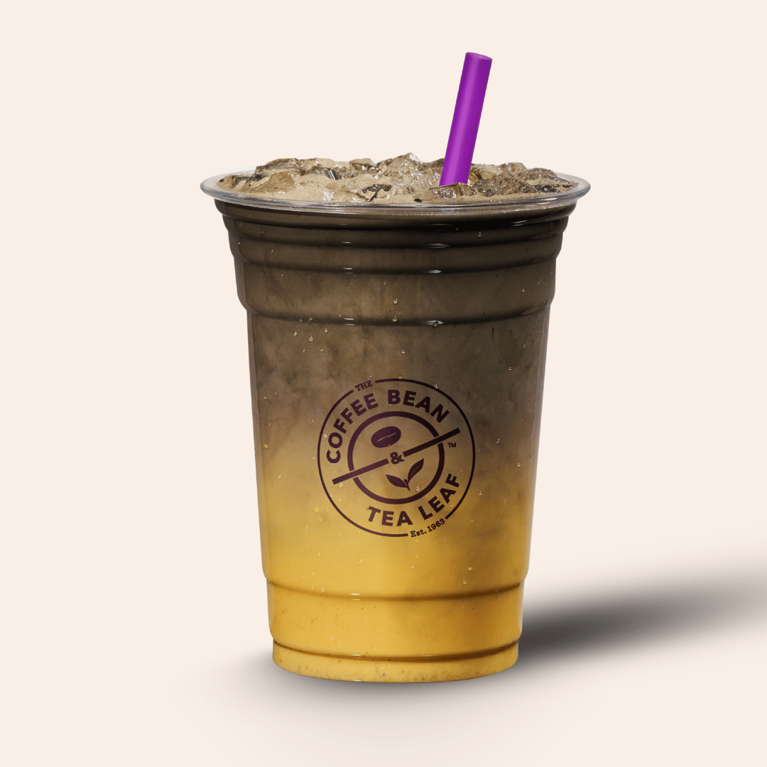 An iced coffee drink in a clear plastic cup with a purple straw, shown on a plain light beige background. The cup has a dark brown upper section and a golden yellow lower section, with the Coffee Bean & Tea Leaf logo printed on the front.