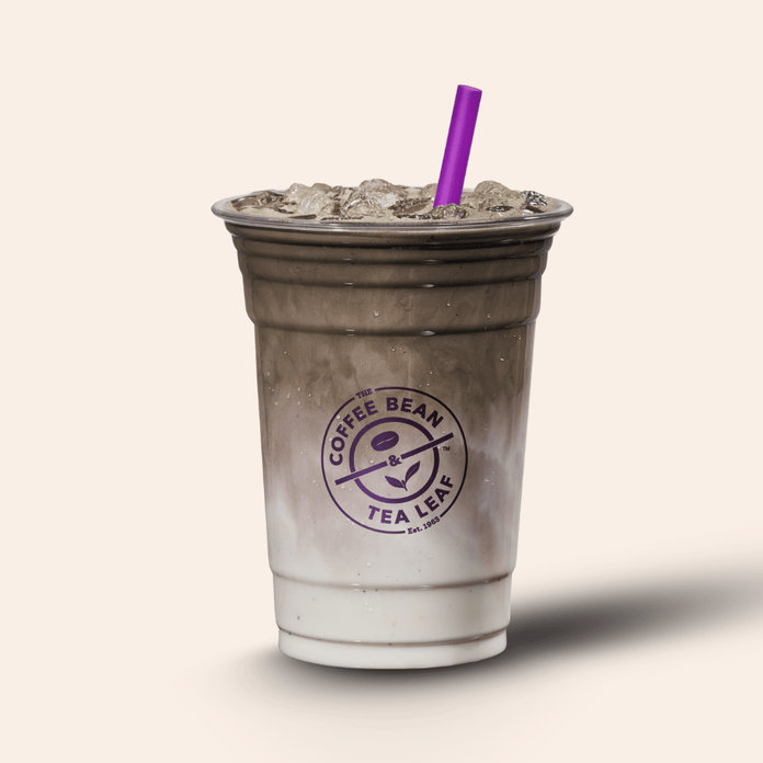 A plastic cup filled with an iced coffee drink and topped with crushed ice, featuring a purple straw and a purple Coffee Bean & Tea Leaf logo on the front, set against a solid light beige background.