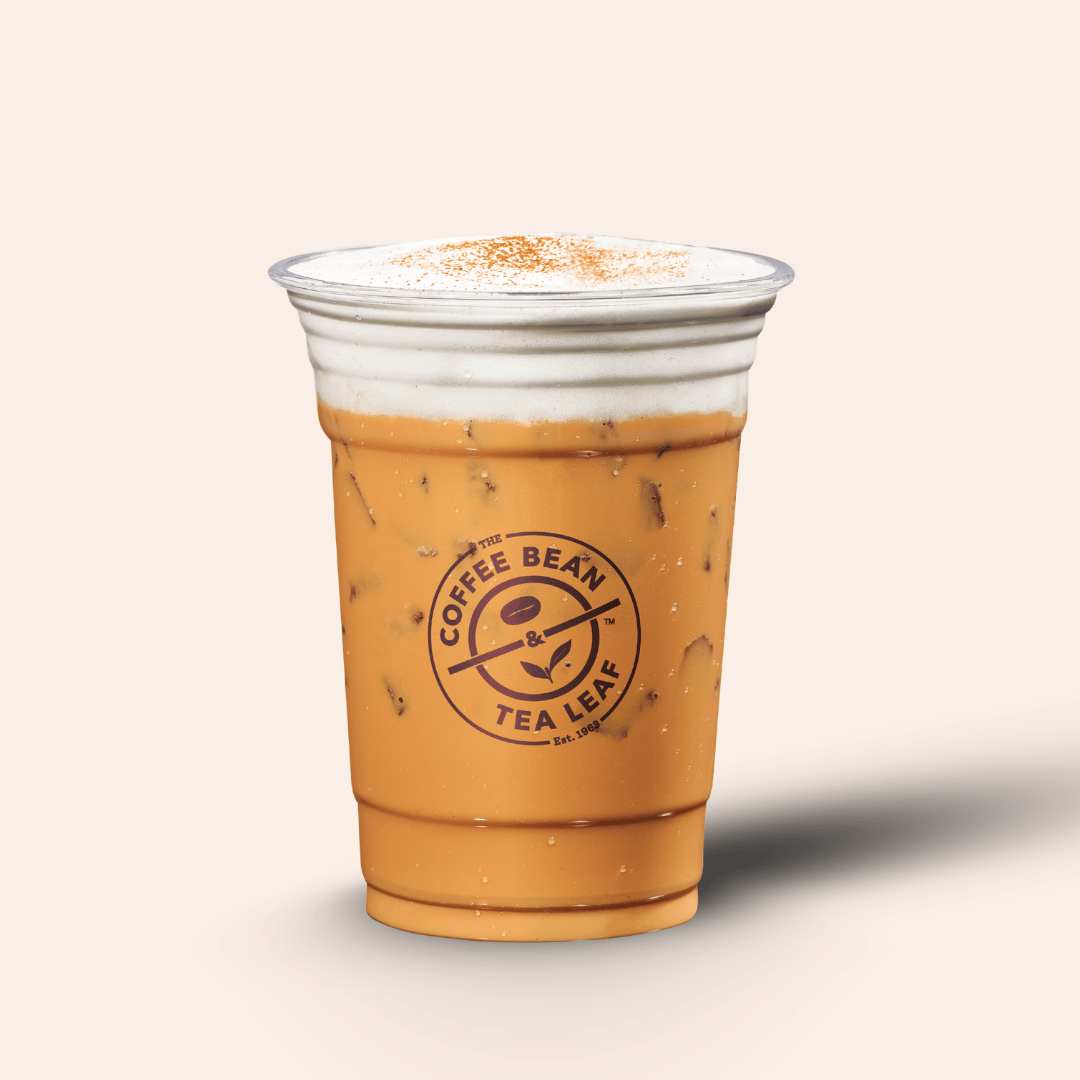 A clear plastic cup of iced coffee or latte with a light brown drink, topped with white foam and a dusting of cinnamon, featuring the Coffee Bean Tea Leaf logo on the front, set against a solid light beige background.