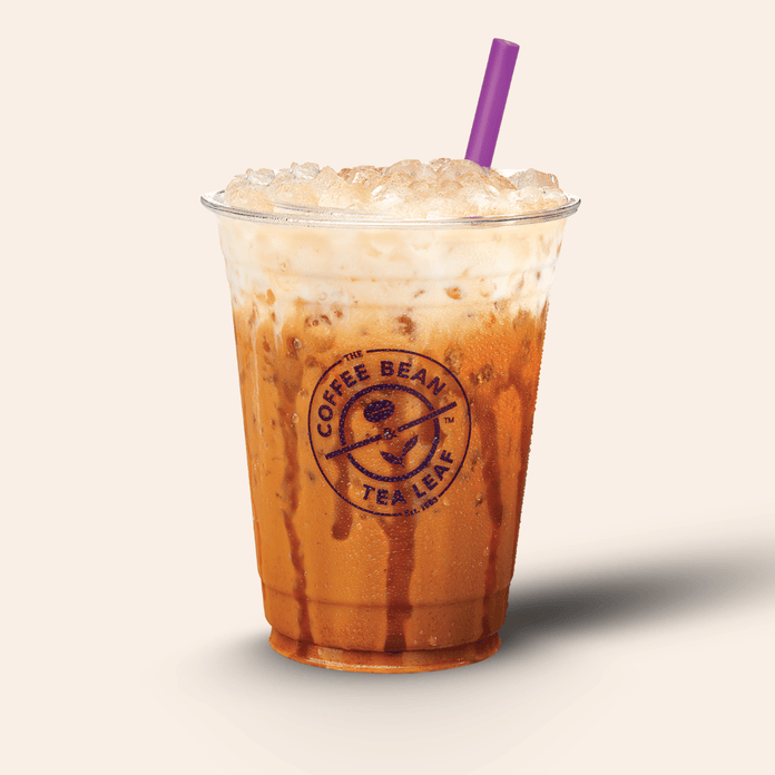 A clear plastic cup filled with iced coffee or latte, topped with whipped cream and crushed ice, with a purple straw. The cup has a visible Coffee Bean Tea Leaf logo on the front, set against a plain light beige background.