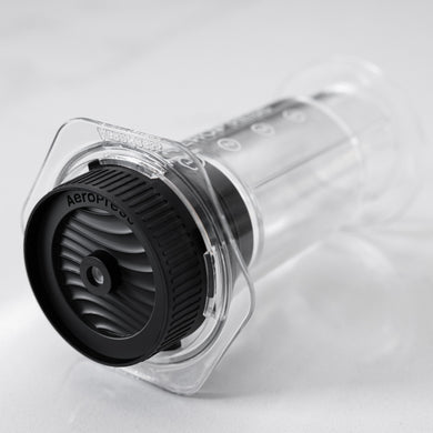 Clear AeroPress coffee brewing device with black AeroPress cap on a light gray background