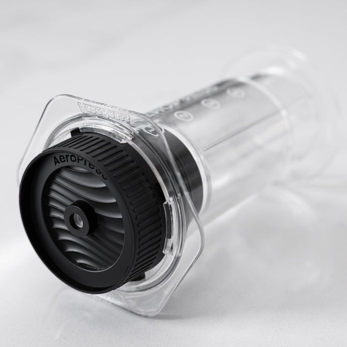 Clear AeroPress coffee brewing device with black AeroPress cap on a light gray background